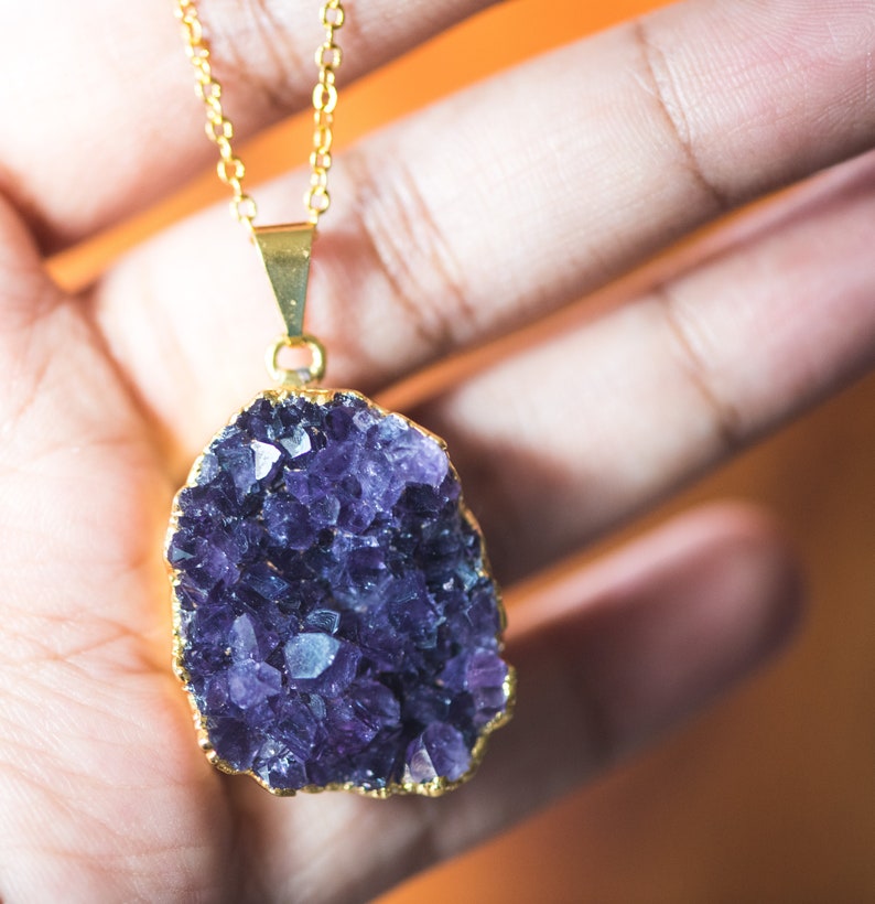 May include: A gold chain necklace with a large amethyst crystal pendant. The crystal is a cluster of purple crystals with a gold rim.
