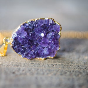 May include: A gold chain necklace with a purple amethyst crystal pendant. The crystal is a cluster of small, faceted stones.
