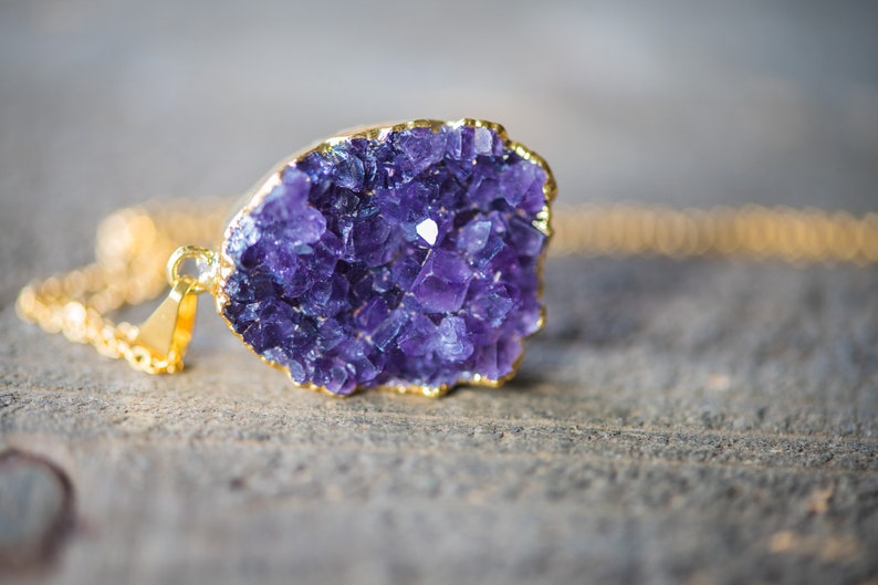 May include: A gold chain necklace with a purple amethyst crystal pendant. The crystal is a cluster of small, faceted stones.