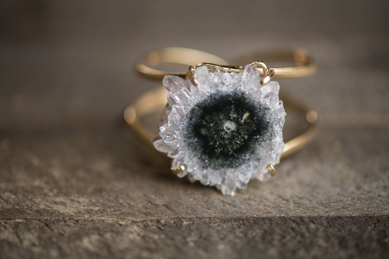May include: A gold-toned cuff bracelet featuring a large, natural geode centerpiece. The geode has a central dark green core surrounded by a ring of light gray and clear crystals. The bracelet's design is simple, highlighting the geode's natural beauty.
