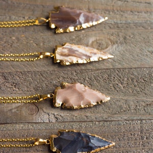 May include: Five gold arrowhead necklaces with different colored stones. The arrowheads are made of agate, jasper, and obsidian. The necklaces are on a wooden surface.
