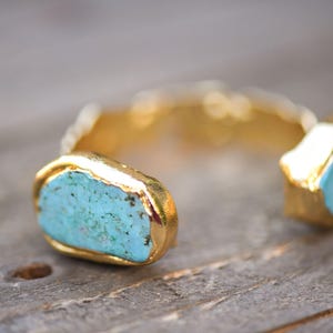 May include: A gold cuff bracelet with two turquoise stones set in the metal. The stones are irregular in shape and have a rough texture.