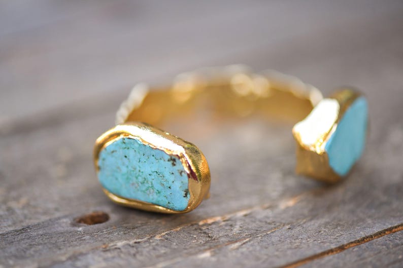 May include: A gold cuff bracelet with two turquoise stones set in the metal. The stones are irregular in shape and have a rough texture.