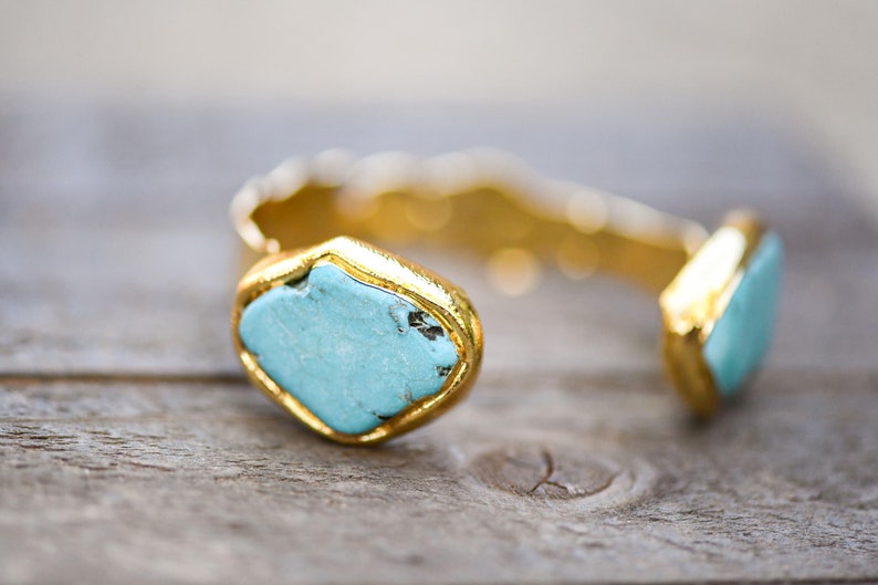 May include: A gold cuff bracelet with two turquoise stones set in the metal. The stones are a light blue color and have a natural, irregular shape.