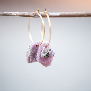 Peut inclure: Une paire de boucles d'oreilles cr&eacute;oles en or avec des pierres de cristal roses et blanches qui pendent. Les boucles d'oreilles sont suspendues &agrave; une branche de bois.