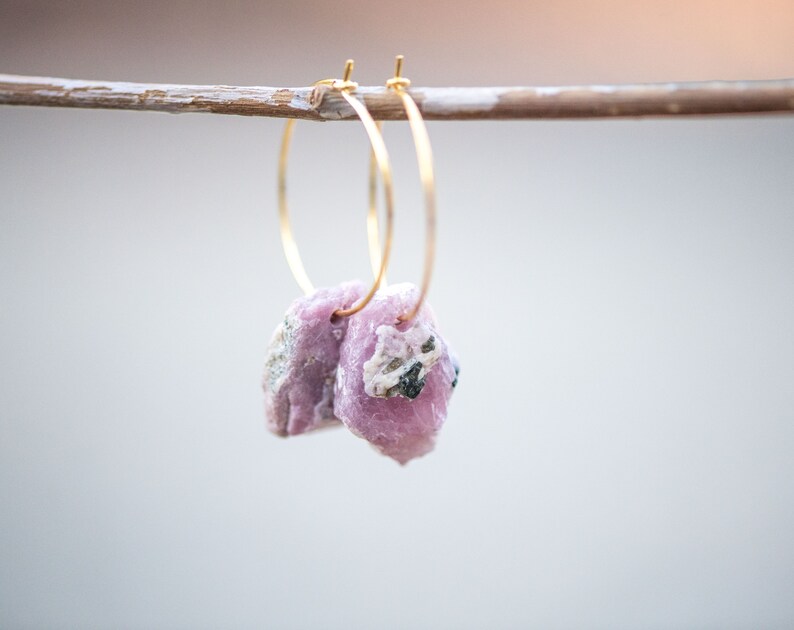 Peut inclure: Une paire de boucles d'oreilles cr&eacute;oles en or avec des pierres de cristal roses et blanches qui pendent. Les boucles d'oreilles sont suspendues &agrave; une branche de bois.