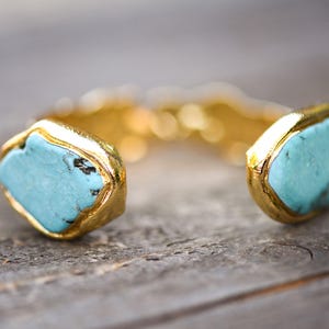May include: A gold cuff bracelet with two turquoise stones set in the center. The stones are a light blue color and have a slightly irregular shape.