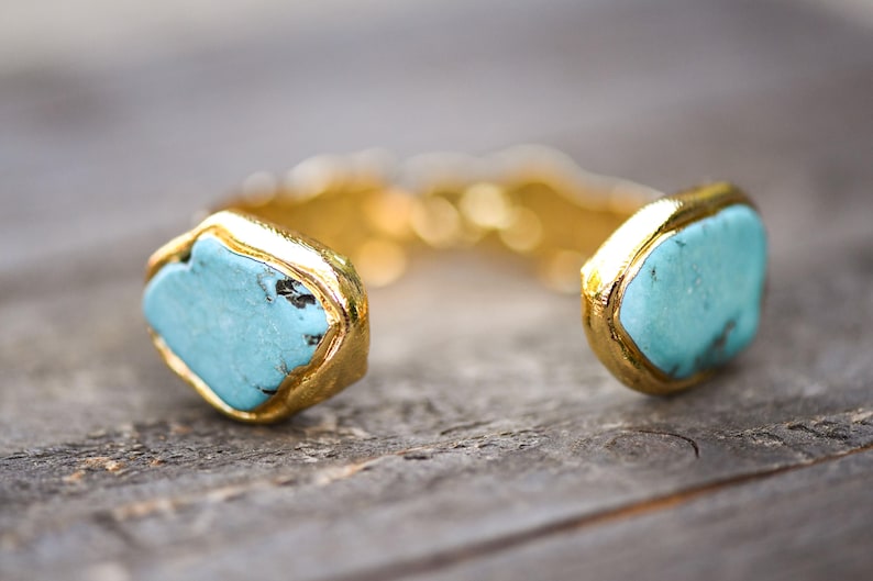 May include: A gold cuff bracelet with two turquoise stones set in the center. The stones are a light blue color and have a slightly irregular shape.