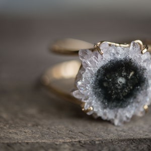 May include: A gold-toned ring featuring a large, natural geode stone. The geode has a central dark area surrounded by a ring of white and clear crystals. The ring's band has a double-loop design.