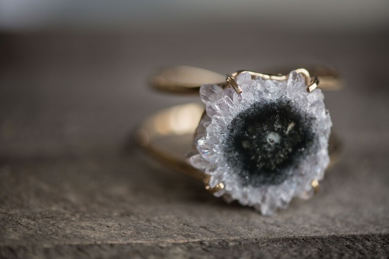 May include: A gold-toned ring featuring a large, natural geode stone. The geode has a central dark area surrounded by a ring of white and clear crystals. The ring's band has a double-loop design.