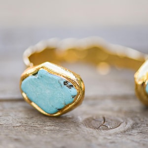 May include: A gold cuff bracelet with two turquoise stones set in the metal. The stones are a light blue color and have a slightly irregular shape.