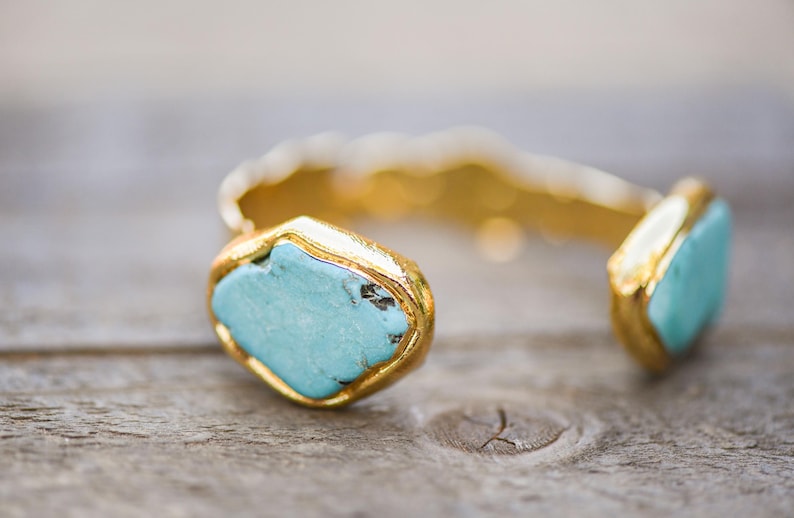May include: A gold cuff bracelet with two turquoise stones set in the metal. The stones are a light blue color and have a slightly irregular shape.