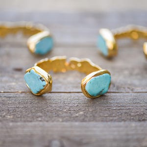 May include: Three gold cuff bracelets with turquoise stones. The bracelets are on a wooden surface.