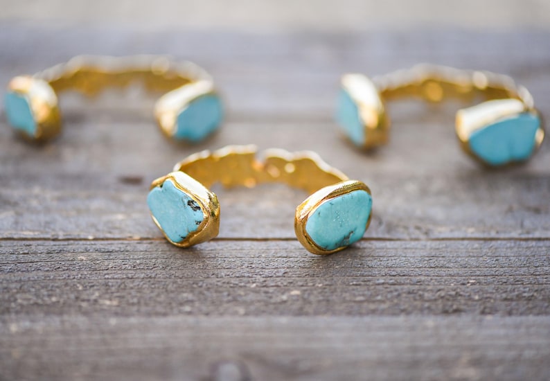May include: Three gold cuff bracelets with turquoise stones. The bracelets are on a wooden surface.