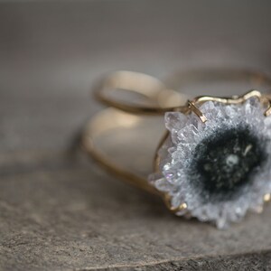 May include: A gold-toned ring featuring a large, round, geode stone with a central dark gray core and a surrounding of light gray crystals. The ring's band is thin and delicate, with a textured surface.