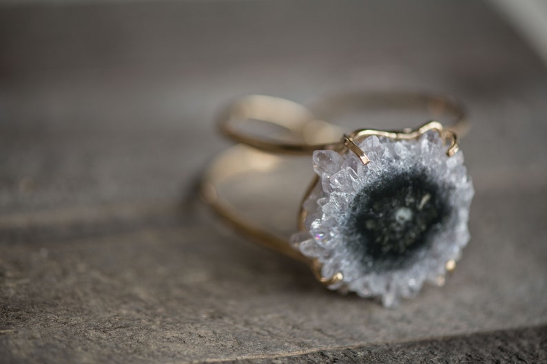 May include: A gold-toned ring featuring a large, round, geode stone with a central dark gray core and a surrounding of light gray crystals. The ring's band is thin and delicate, with a textured surface.