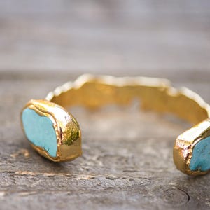 May include: A gold cuff bracelet with two turquoise blue stones set into the metal. The bracelet is open and ready to be worn.
