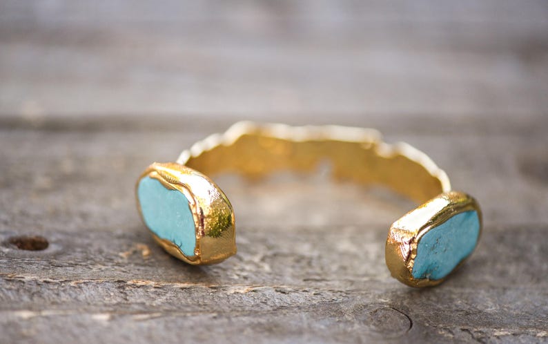 May include: A gold cuff bracelet with two turquoise blue stones set into the metal. The bracelet is open and ready to be worn.