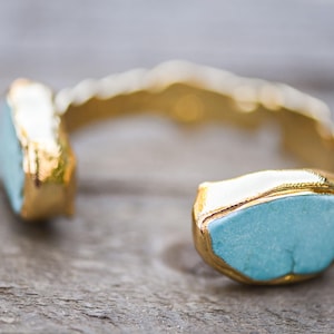 May include: A gold cuff bracelet with two turquoise blue stones. The stones are set in gold and have a rough, natural look.