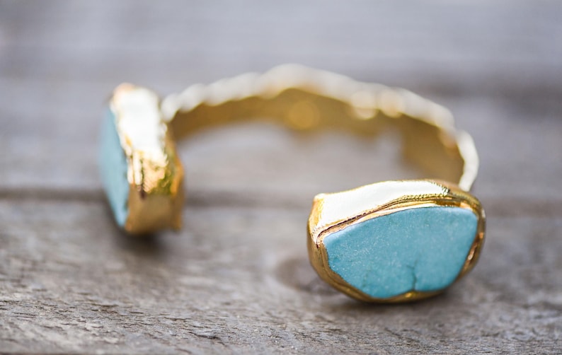 May include: A gold cuff bracelet with two turquoise blue stones. The stones are set in gold and have a rough, natural look.