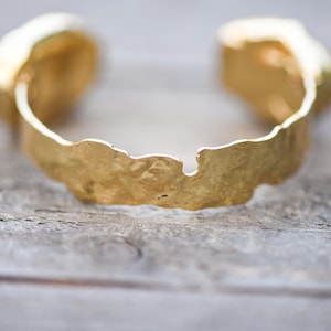 May include: A gold-toned bracelet with a textured, irregular design. The bracelet is open and has a slightly curved shape.