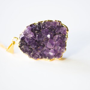 May include: A gold-plated pendant featuring a large amethyst crystal cluster. The crystal is a deep purple color and has a natural, rough texture. The pendant is perfect for adding a touch of elegance and natural beauty to any outfit.