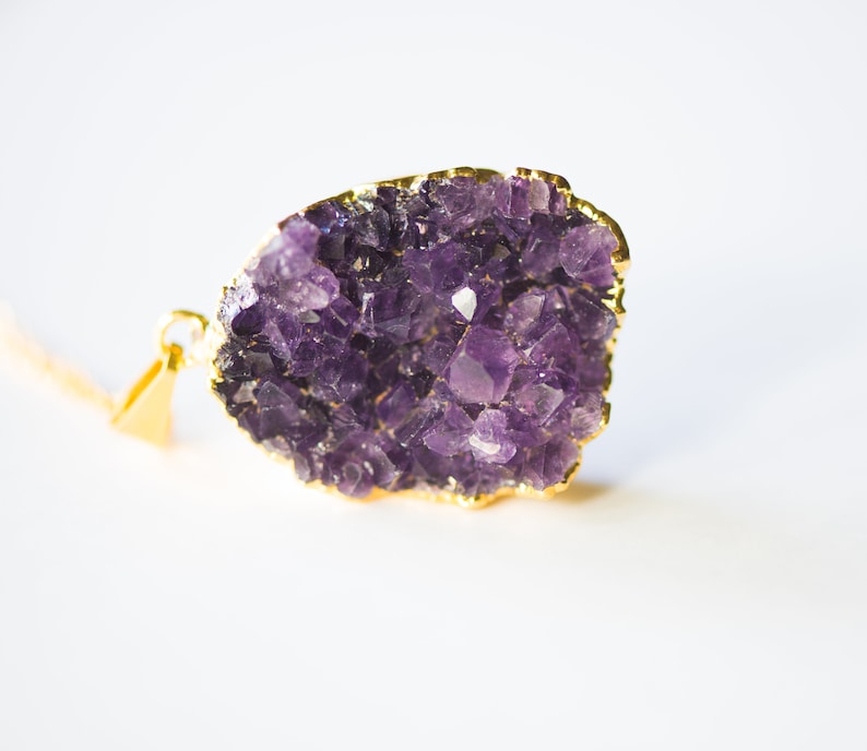 May include: A gold-plated pendant featuring a large amethyst crystal cluster. The crystal is a deep purple color and has a natural, rough texture. The pendant is perfect for adding a touch of elegance and natural beauty to any outfit.