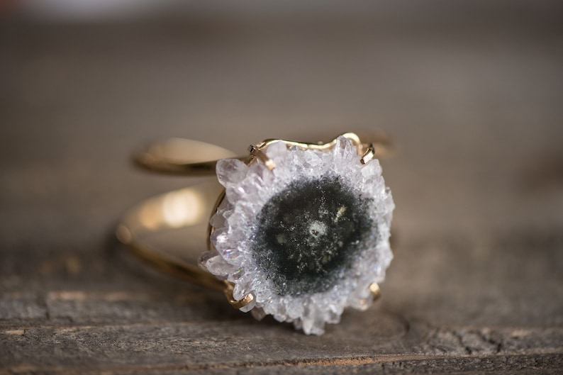 May include: A gold-toned ring featuring a central, circular stone with a dark center and crystalline, light-colored edges. The ring's band splits into two parallel lines, creating a unique design. The ring is set against a blurred wooden background.