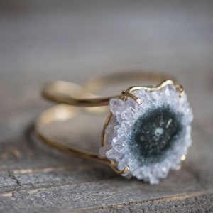May include: A gold-toned bracelet featuring a central, round, geode stone with a dark center and crystalline edges. The bracelet has a double-band design, showcasing the natural beauty of the gemstone. The background is a wooden surface.