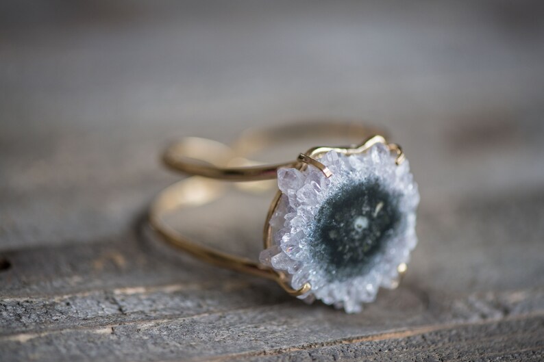 May include: A gold-toned bracelet featuring a central, round, geode stone with a dark center and crystalline edges. The bracelet has a double-band design, showcasing the natural beauty of the gemstone. The background is a wooden surface.
