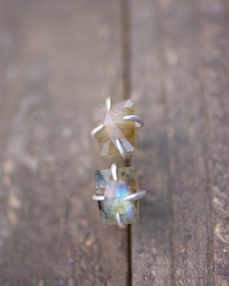 Pu&ograve; includere: Un paio di orecchini a lobo in argento con pietre preziose di labradorite iridescente. Le pietre sono incastonate in una montatura a griffe e hanno una forma rettangolare.