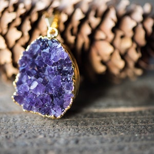 May include: A gold pendant featuring a large amethyst crystal with a cluster of purple crystals. The pendant is on a wooden surface.