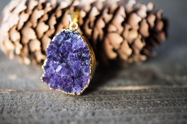 May include: A gold pendant featuring a large amethyst crystal with a cluster of purple crystals. The pendant is on a wooden surface.