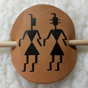 May include: A brown leather hair stick with two black painted figures on it. The figures are stylized and have a geometric design. The stick is about 4 inches long.