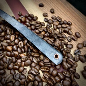 May include: A close-up of a dark gray metal coffee scoop with a copper rivet. The scoop is surrounded by coffee beans on a wooden surface.