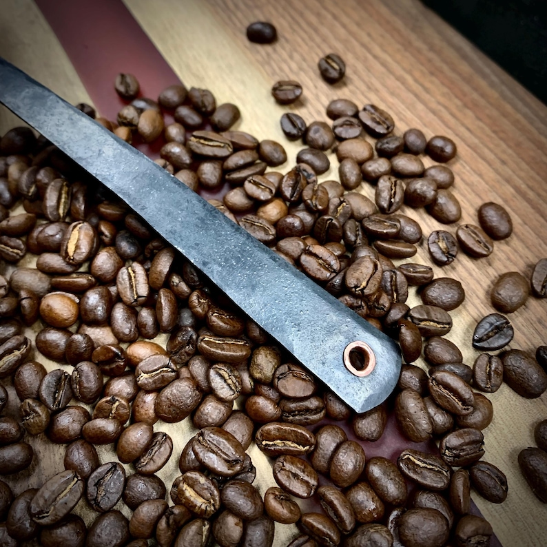 May include: A close-up of a dark gray metal coffee scoop with a copper rivet. The scoop is surrounded by coffee beans on a wooden surface.