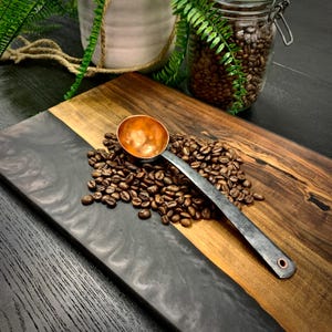 May include: A copper and black metal scoop sits on a wooden cutting board with a black epoxy resin inlay. The scoop is surrounded by coffee beans.