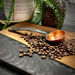 May include: A wooden cutting board with a black epoxy resin inlay. A copper scoop sits on a pile of coffee beans on the board.