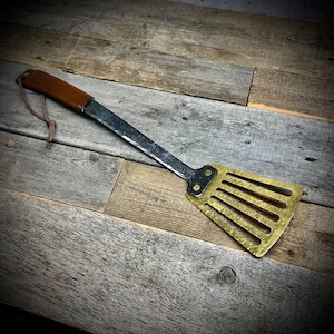 May include: A long-handled spatula with a brown leather handle and a brass blade with a hammered finish. The blade has a series of slots for flipping food.