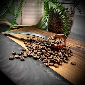 May include: A copper scoop filled with roasted coffee beans sits on a wooden cutting board with a black and gray epoxy resin inlay. The scoop is made of hammered metal and has a long, curved handle.