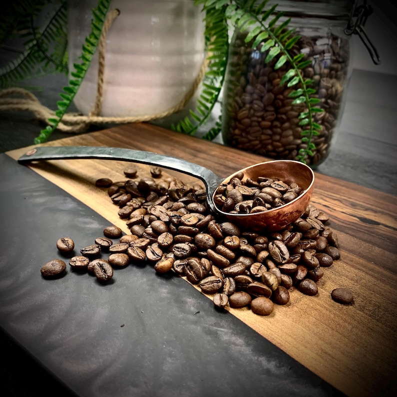 May include: A copper scoop filled with roasted coffee beans sits on a wooden cutting board with a black and gray epoxy resin inlay. The scoop is made of hammered metal and has a long, curved handle.