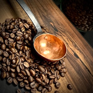 May include: A copper coffee scoop with a black metal handle. The scoop is sitting on a pile of coffee beans.