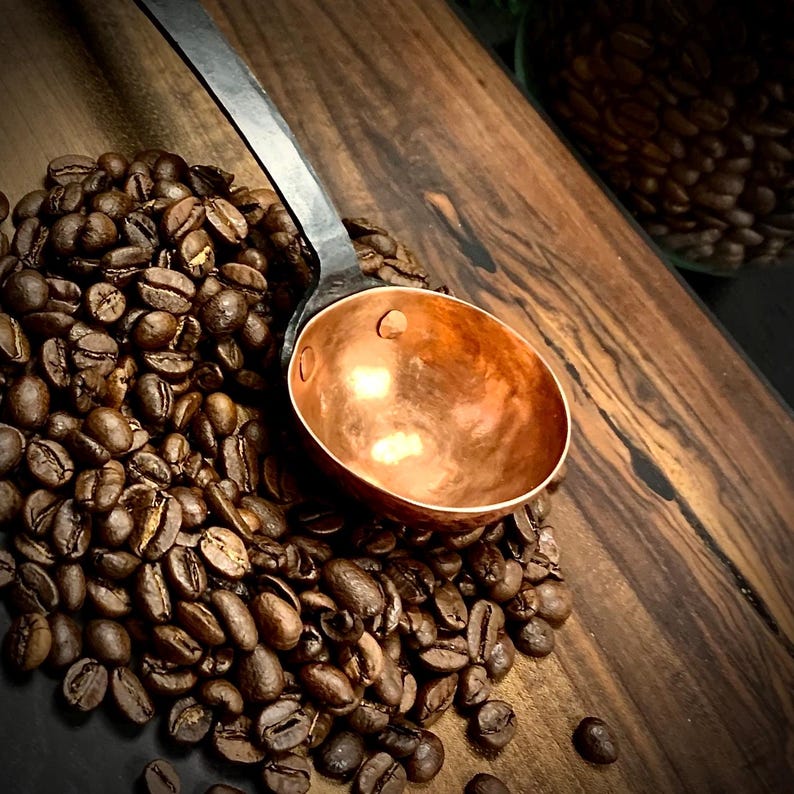 May include: A copper coffee scoop with a black metal handle. The scoop is sitting on a pile of coffee beans.