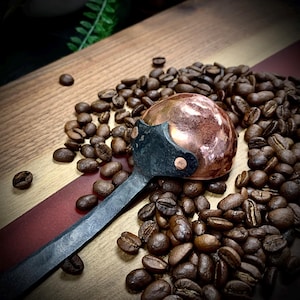 May include: A copper and black coffee scoop with a rounded bowl and a long handle. The scoop is sitting on a wooden surface surrounded by coffee beans.