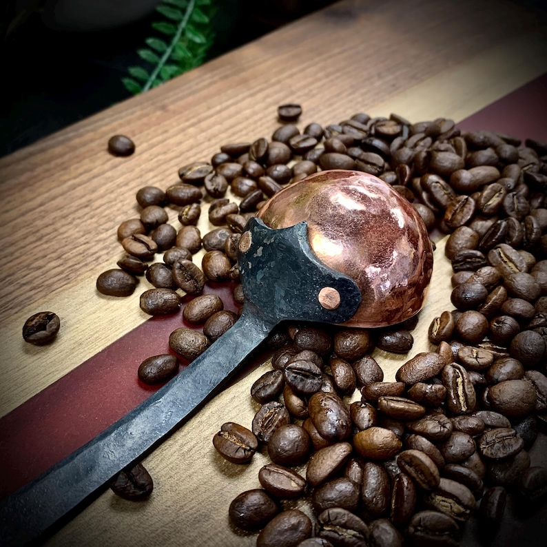 May include: A copper and black coffee scoop with a rounded bowl and a long handle. The scoop is sitting on a wooden surface surrounded by coffee beans.