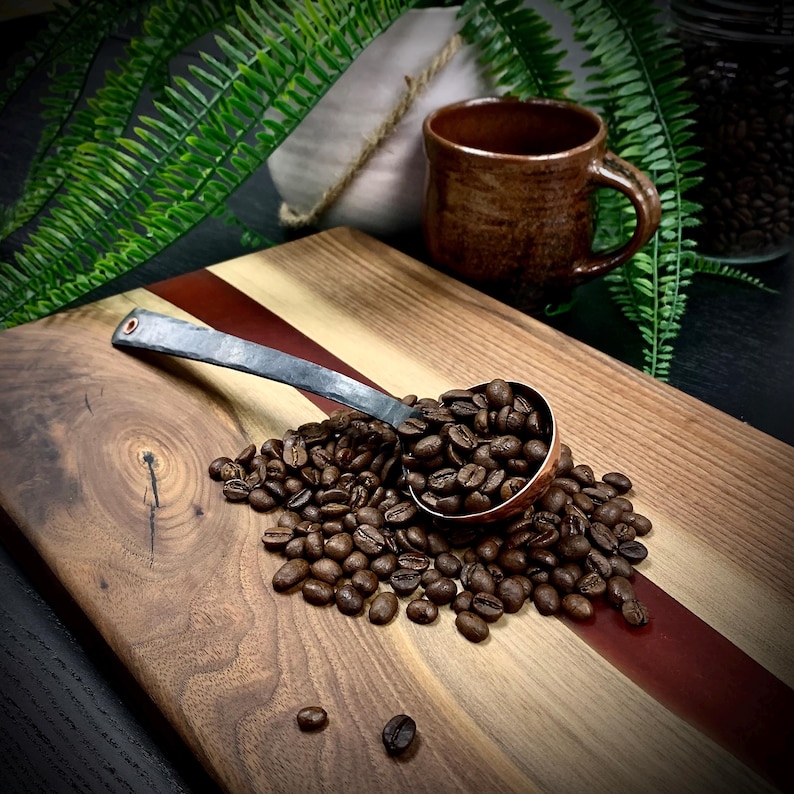 May include: A wooden cutting board with a red stripe and a copper scoop filled with coffee beans. The scoop is made of metal with a black handle.