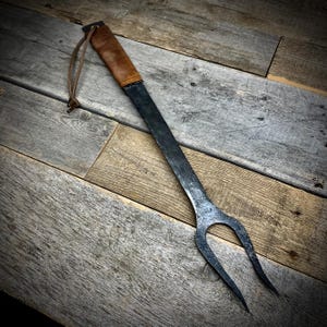 May include: A long, black metal grilling fork with a brown leather handle and a leather strap. The fork has two tines and is resting on a wooden surface.