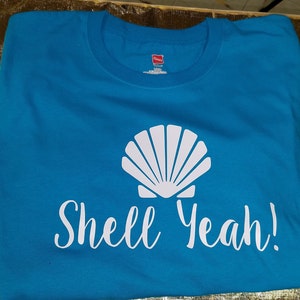 May include: A turquoise t-shirt with a white seashell graphic and the phrase "Shell Yeah!" printed in white script. The shirt is made of a soft material and is suitable for casual wear.