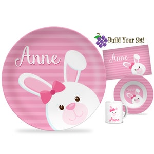 May include: Pink and white personalized children's dinnerware set with a bunny rabbit design. The set includes a plate, bowl, mug, and placemat. The name "Anne" is printed on each piece.