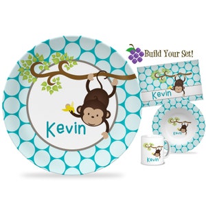 May include: A children's dinnerware set featuring a cartoon monkey hanging from a tree branch, holding a banana. The set includes a plate, bowl, placemat, and mug, all with a teal polka dot pattern and the name "Kevin".